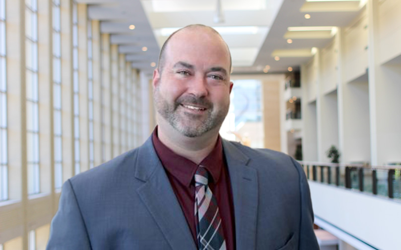 Headshot of Brad Clinton wearing a gray blazer, maroon shirt, and striped tie standing on an indoor balcony overlooking large windows and a sculpture in background.