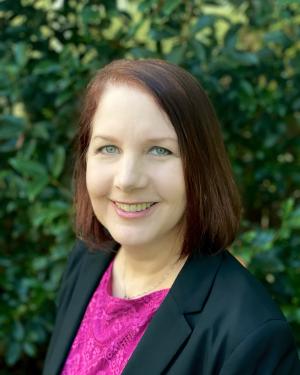 Headshot of Dawn Delventhal standing in front of shrubbery while smiling and wearing a pink top and black blazer.