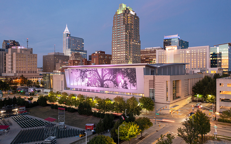 The Downtown Raleigh skyline at dusk, with the convention center and its illuminated shimmer wall featured prominently in the center.