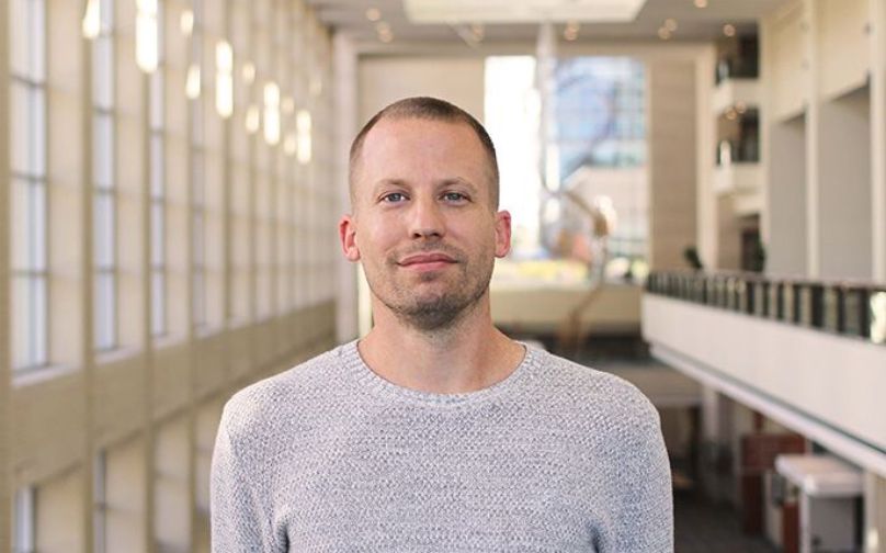 Headshot of John Hughes wearing a light gray top standing on an indoor balcony overlooking large windows and a sculpture in background.