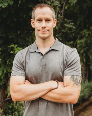 Headshot of Jon Curran wearing a gray polo and standing in front of shrubbery with arms crossed.