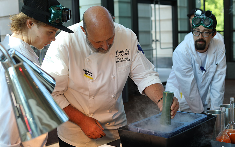 A chef prepares a dish with two people watching.