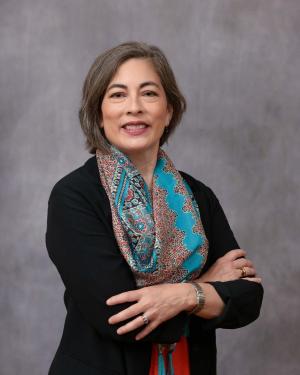 Headshot of Wendy-Jo Toyoma standing in front of gray backdrop with arms crossed wearing a black top and blue scarf.