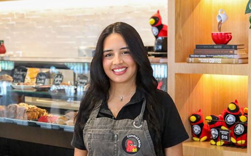 Headshot of Zikyu wearing a black polo and Complex Brew branded apron standing in front of the baked goods counter and shelves with books and cardinal plushies.