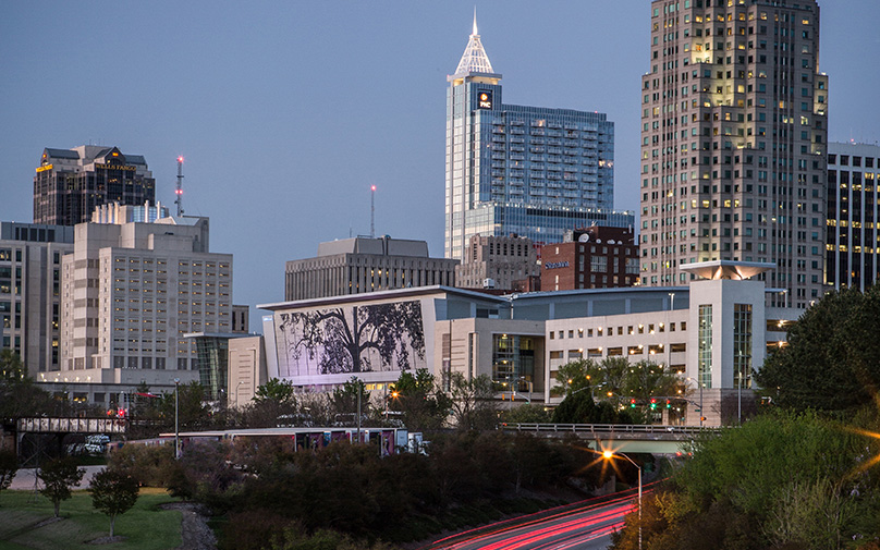 View of Atlantic Union Bank Convention Center shimmer wall with downtown Raleigh buildings in the back and trees in front.