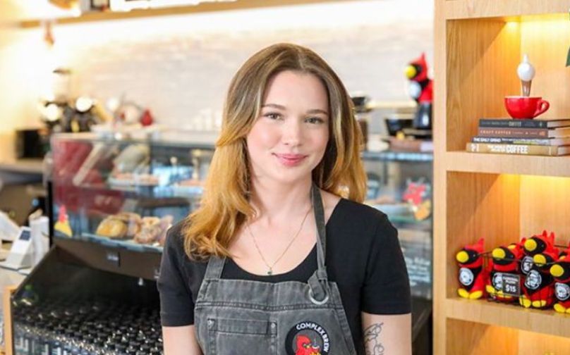 Headshot of Jess wearing a black polo and Complex Brew branded apron standing in front of the baked goods counter and shelves with books and cardinal plushies.