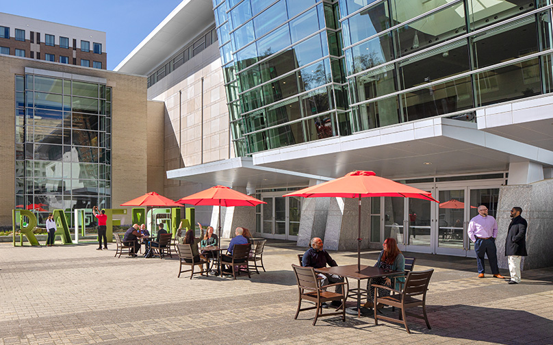 View of the entrance to Atlantic Union Bank Convention Center from the side with people gathered and sitting at tables with red umbrellas..